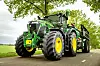 Grønn John Deere-traktor med gulhjul og tilhenger på en landevei under trær.