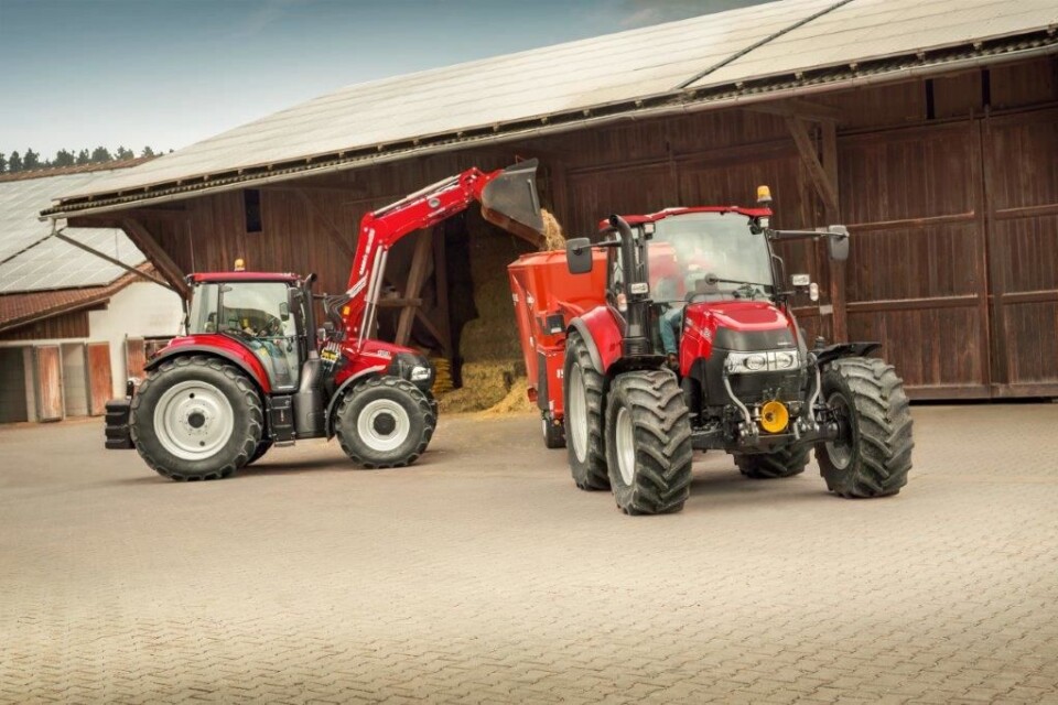 Case IH Luxxum fyller fôr i ei fôrvogn trukket av en annen Case IH-traktor foran brune driftsbygninger.