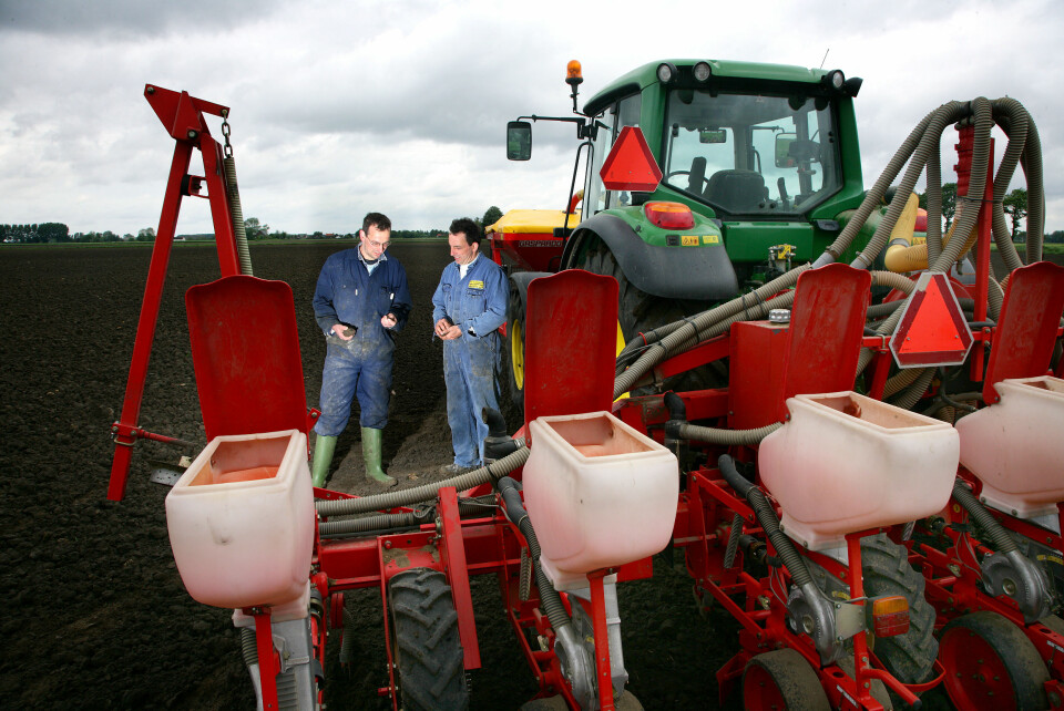 To bønder står og diskuterer noe på jordet ved siden av en John Deere traktor med såmaskin bak.