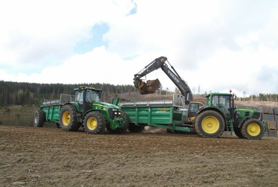 To John Deere traktorer med hver sin henger som blir fylt opp med en gravemaskin.