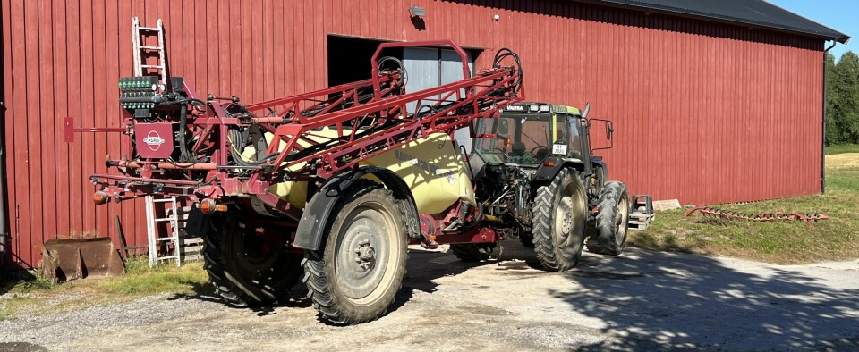 Valtra traktor med Hardi åkersprøyte foran en rødmalt driftsbygning.